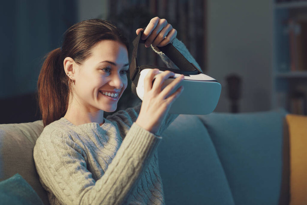 Smiling young woman sitting on the couch at home and wearing a VR headset, entertainment and virtual reality concept - Foto, Imagem