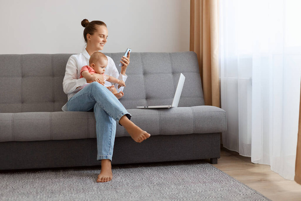 Portrait of happy woman in white t shirt sitting on sofa with baby daughter and working on laptop, holding smart phone in hands, searching information or chatting with friends. - Fotoğraf, Görsel