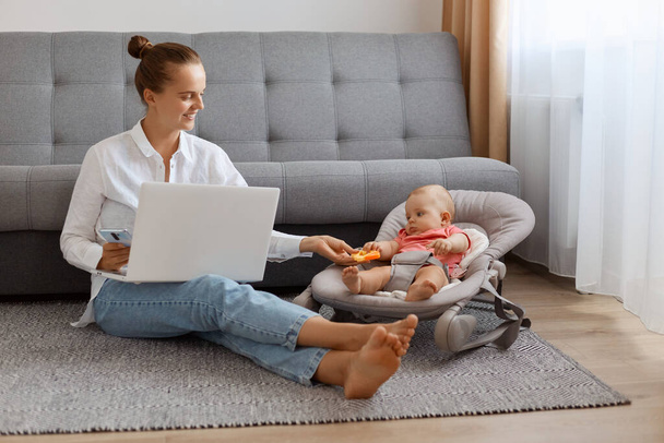 Image of delighted busy woman in white t shirt sitting near cough with baby daughter in rocking chair and working on laptop, typing on notebook and showing toy to her kid. - 写真・画像