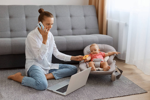 Indoor shot of attractive woman in white t shirt sitting near cough with baby daughter in rocking chair and working on laptop, talking on phone with business partner. - Valokuva, kuva
