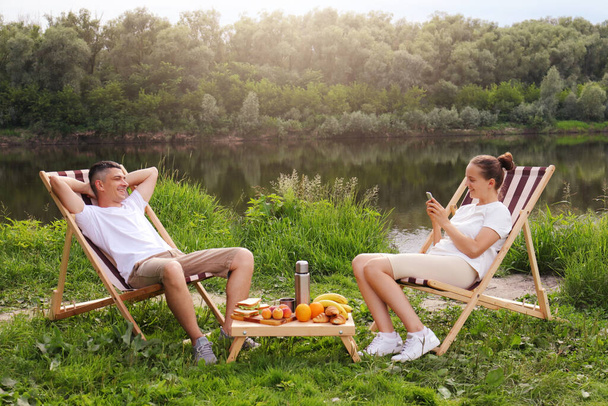 Outdoor shot of young couple sitting on deck chair and talking near the river, relaxed man sits with hands behind head, woman using cell phone for checking social networks, family relaxing outdoor. - Foto, imagen