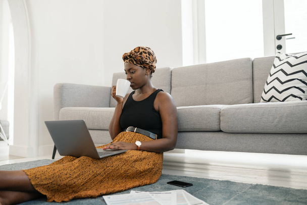 Beautiful African woman sitting on floor in lounge, work meeting on laptop at home with coffee in hand wearing traditional head scarf, sipping on cup of coffee - Photo, image