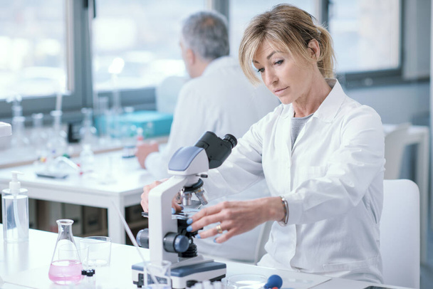 Female medical researcher working in the lab, she is sitting at desk and using a microscope - Foto, Bild