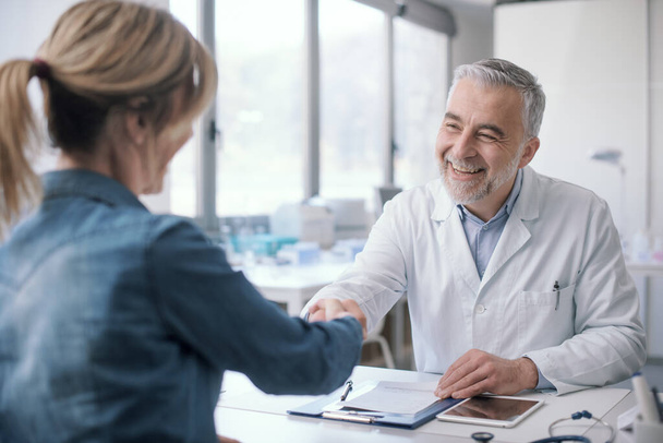 Smiling doctor sitting at desk and giving a handshake to his patient, medicine and healthcare concept - Foto, Bild