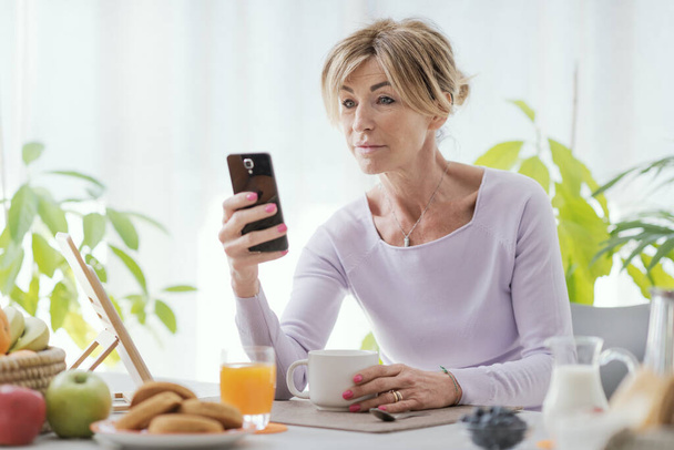 Mature woman having breakfast and connecting with her smartphone at home - Foto, Imagen