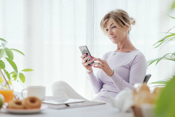 Mature woman having breakfast and connecting with her smartphone at home - Foto, Imagem