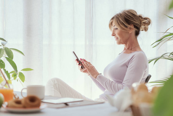 Mature woman having breakfast and connecting with her smartphone at home - Photo, image