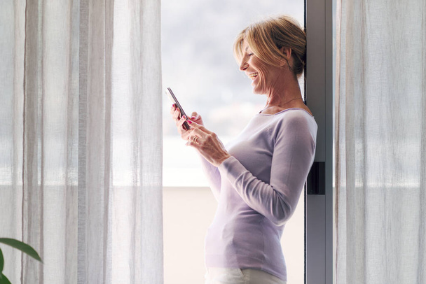 Happy mature woman standing next to a window at home and connecting with her smartphone - Photo, image