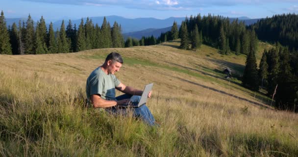 Чоловік сидить на красивому лузі в горах, працює на ноутбуці. Концепція фрілансу, цифрової кочівниці або віддаленого офісу
. - Кадри, відео