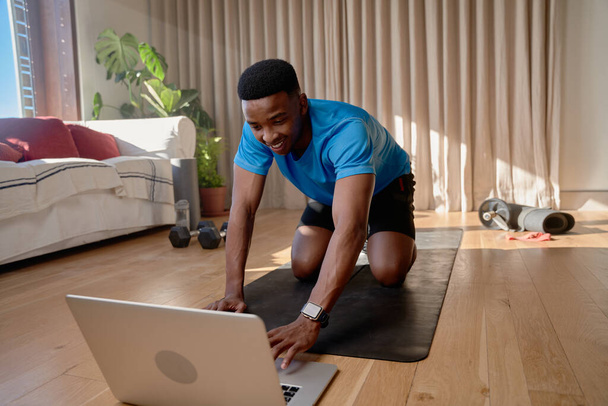 Young happy African america male working out at home doing an online class using laptop watching videos on an exercise mat in his living room. High quality photo - Foto, Bild
