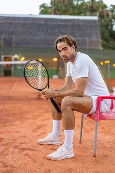 Caucasian blonde long haired young man sitting on a chair holding tennis racket on a dirt court. - Zdjęcie, obraz