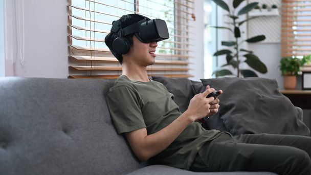 Joyful asian male experiencing opportunities of gaming in virtual reality headset, relaxing on couch at home. - Foto, immagini