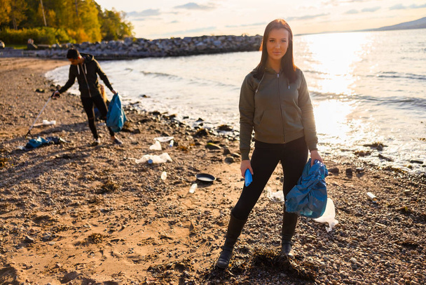 Портрет серйозної молодої жінки, яка тримає мішок для сміття. Добровольці прибирають пляж для пластику проти неба під час прекрасного заходу сонця. Вони піклуються про екологічні збитки
. - Фото, зображення