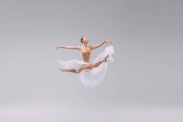 Портрет молодой балерины, танцующей со светлой прозрачной тканью на сером фоне студии. Летает как птица. Свободу. Концепция классического балета, вдохновения, красоты, танца, творчества - Фото, изображение