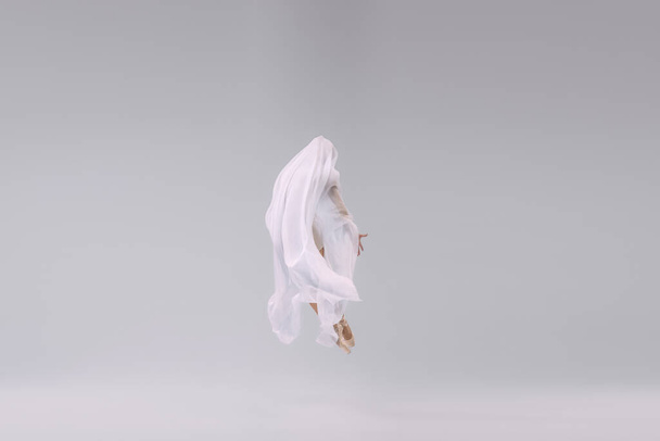 Portrait of young ballerina dancing, jumping fully covered with transparent fabric isolated over grey studio background. Concept of classic ballet, inspiration, beauty, dance, creativity - Fotó, kép