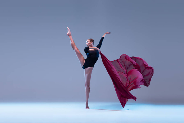 Портрет молодої гнучкої балерини, що танцює з глибокою червоною тканиною, ізольованою на синьо-сірому студійному фоні. Вишуканий. Концепція класичного балету, натхнення, краси, танцю, творчості
 - Фото, зображення