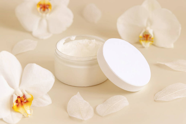Opened Cream jar with a blank white lid near white orchid flowers on light yellow close up. Cosmetic packaging Mockup. Exotic natural cosmetics, romantic composition, everyday skincare routin - Foto, Imagem