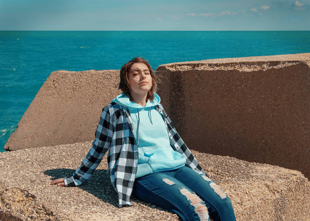 Relaxed Teenager girl in light blue hoodie and plaid shirt. Smiling Teen girl sitting outdoor and enjoying the sunny day. Adolescence concept - 写真・画像