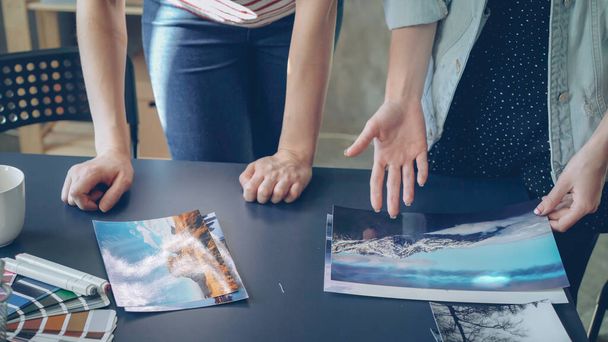 Наклонный снимок женских рук, размещающих фотографии на современном столе в современном офисе. Женщины-дизайнеры группируют картинки по цвету и изображению и обсуждают иллюстрации
. - Фото, изображение