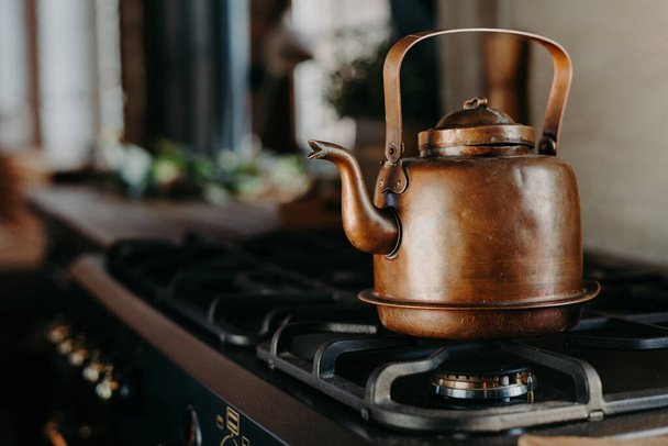 Bronze kettle in modern kitchen. Old vintage teapot on gas stove. Preparing tea. Aluminium teakettle. Sunny daylight from window. - Photo, image