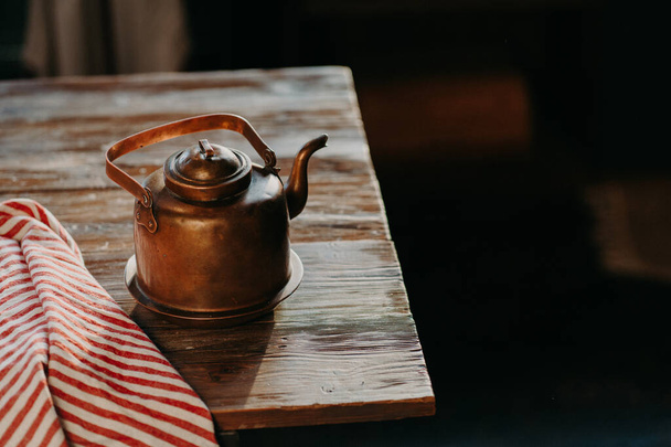 Παλιά τσαγιέρα από χαλκό σε ξύλινο τραπέζι σε σκοτεινό δωμάτιο. Κόκκινη ριγέ πετσέτα κοντά. Βραστήρας για τσάι ή καφέ. Εξοπλισμός μαγειρέματος - Φωτογραφία, εικόνα