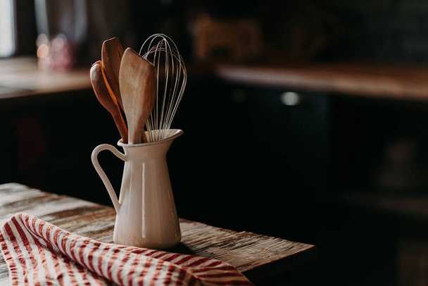 Кухонные принадлежности на деревянном столе. Посуда в белой керамической банке на темном фоне. Сельский стиль. Посуда для приготовления еды. Деревянные ложки шпатула и венчик - Фото, изображение