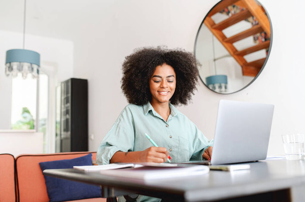 Positive smart young multiracial smiling female student sitting at the table with laptop, studying from home and taking notes to the notebook, watching an online webinar or taking a course - Fotó, kép