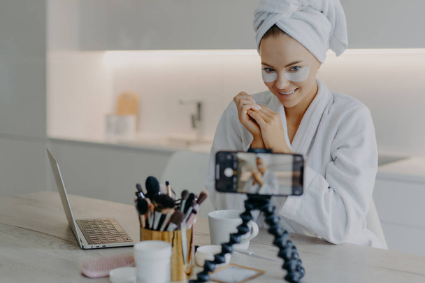 Pleased beauty blogger sits in front of cellphone camera shares experience about skin care wears hydrogel patches under eyes poses at table with cosmetic tools wears bathrobe wrapped towel on head - Photo, Image