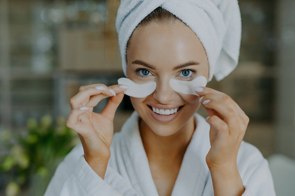 Headshot of happy young blue eyed woman with minimal makeup takes off beauty patches enjoys good effect has healthy well cared skin white perfect teeth wears bathrobe and wrapped towel on head - Photo, Image
