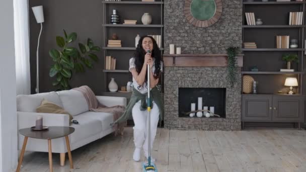 Счастливая юная смешная женщина убирает в современной гостиной поет песню, используя швабру в качестве микрофона активной энергичной домохозяйки наслаждаясь уборка моет пол нелепой танцевальной уборки реклама агентства - Кадры, видео