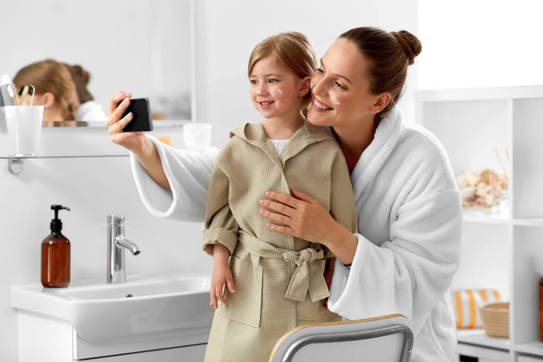 schoonheid, familie en mensen concept - gelukkig glimlachende moeder en dochter met moisturizer op hun gezichten selfie met smartphone in de badkamer - Foto, afbeelding