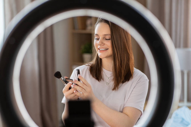 beauty blogging, technology and people concept - happy smiling girl blogger with ring light and smartphone applying make up at home - Photo, Image