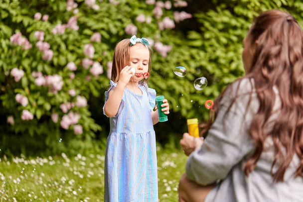 family, motherhood and people concept - happy mother with little daughter blowing soap bubbles at summer park or garden - Foto, Imagem