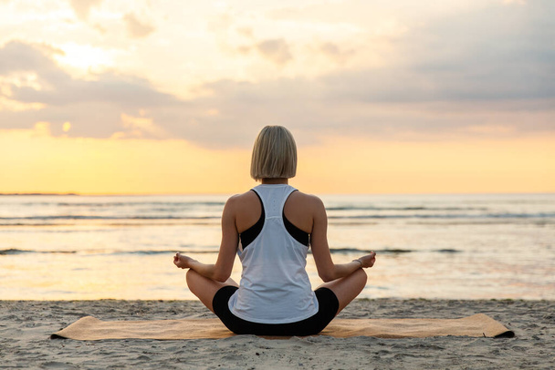 yoga, mindfulness and meditation concept - woman meditating in lotus pose on beach over sunset - Foto, Bild