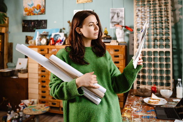 Young white woman wearing sweater examining her drawings in her studio - Фото, зображення