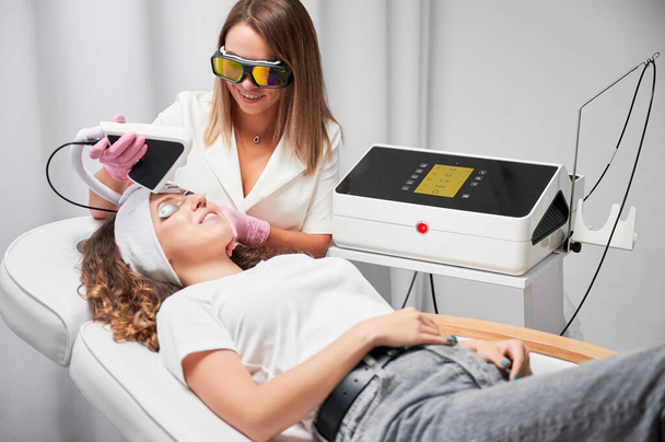 Женщина-косметолог, использующая прибор лазерного сканирования диодов во время повторной процедуры по уходу за кожей. Улыбающаяся женщина в защитных очках получает лазерное лечение лица в салоне красоты. - Фото, изображение