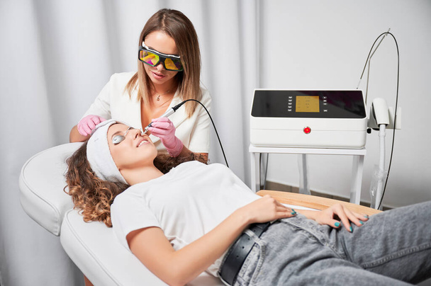 Modern treatment of telangiectasias on skin. Beautician performing local high-precision removal of dilated small vessels on patient face with innovative yellow laser. - Foto, Imagem