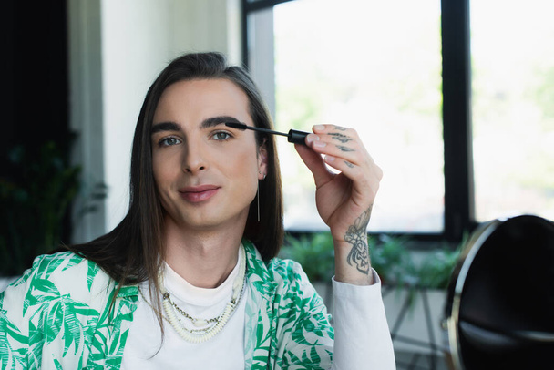 Queer designer applying mascara brush near blurred mirror in office  - Foto, Bild