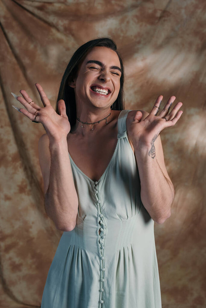 Cheerful tattooed nonbinary person in dress closing eyes on abstract brown background  - Foto, Imagem