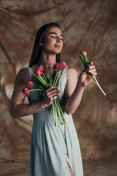 Young nonbinary person in sundress holding pink tulips on abstract brown background - Foto, Imagen