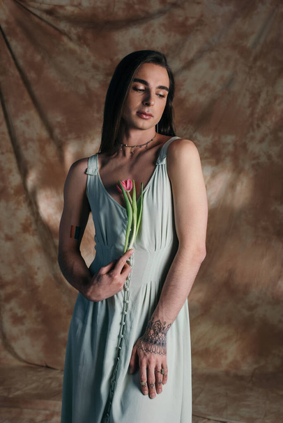 Young brunette queer person in dress holding pink tulip on abstract brown background  - Fotografie, Obrázek