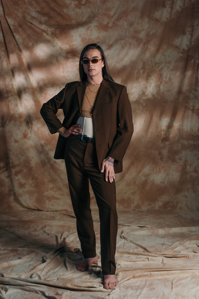 Повна довжина стильної квір-людини в костюмі і корсеті, що стоїть на абстрактному коричневому фоні
 - Фото, зображення