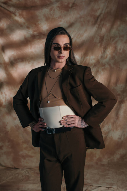 Brunette queer person in sunglasses holding hands on hips on abstract brown background - Fotoğraf, Görsel