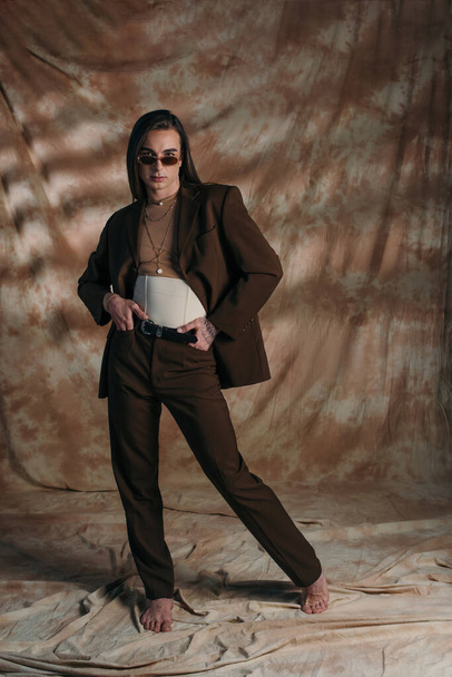 Barefoot queer person in suit and corset posing on abstract brown background - Photo, Image