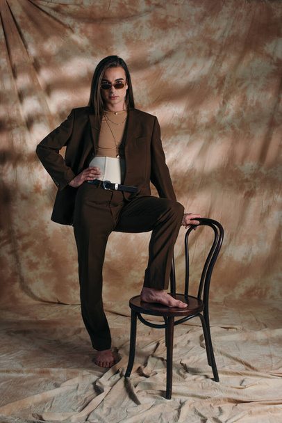 Stylish nonbinary person in suit and sunglasses posing near chair on abstract brown background - Fotografie, Obrázek