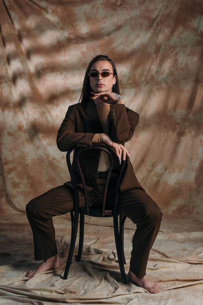 Barefoot queer person in sunglasses and suit sitting on chair on abstract brown background - Φωτογραφία, εικόνα