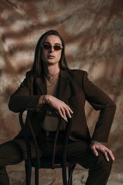 Stylish queer person in suit and sunglasses sitting on chair on abstract brown background - Photo, image