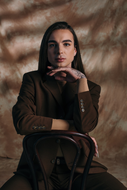 Queer person in jacket looking at camera while sitting on chair on abstract brown background - Foto, Imagen