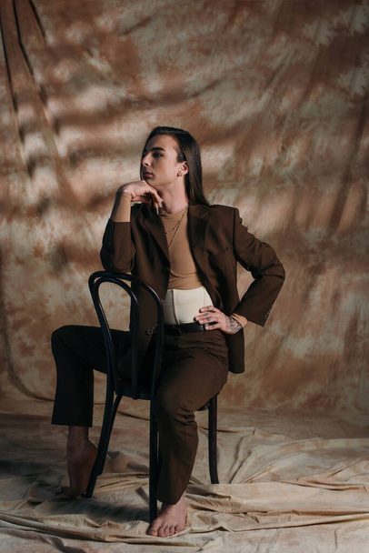 Fashionable queer person in jacket sitting on chair on abstract brown background - Fotoğraf, Görsel