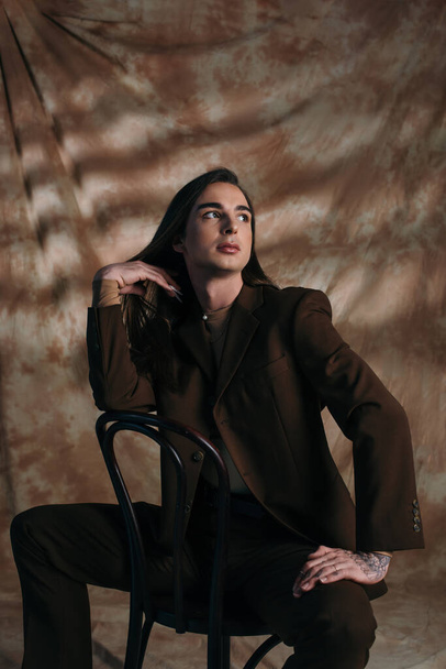 Trendy queer person in suit touching hair while sitting on chair on abstract brown background - Φωτογραφία, εικόνα